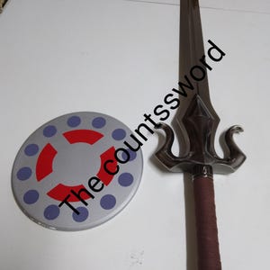 Könnte beinhalten: Ein Schwert- und Schild-Set. Das Schwert hat eine dunkle Klinge und einen braun umwickelten Griff. Der Schild ist rund, silberfarben mit roten und blauen Akzenten. Der Text "The countssword" befindet sich auf dem Bild.