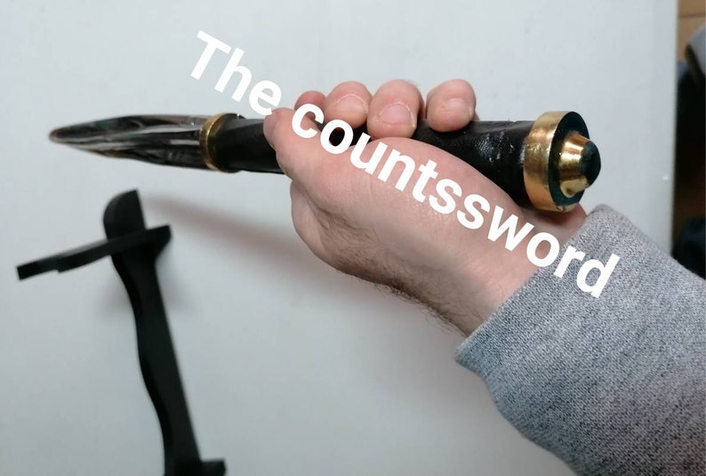 May include: A black and silver dagger with a gold-colored hilt is held in a hand. The dagger has a pointed blade and a black handle. The text "The countssword" is visible in the image.