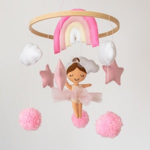 May include: A pink and white baby mobile with a ballerina, stars, clouds, and pom-poms hanging from a wooden hoop. The ballerina is wearing a pink tutu and has brown hair.