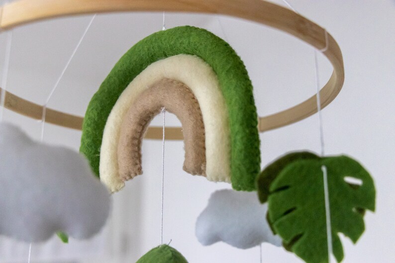 May include: A wooden ring with a green, white, and brown felt rainbow hanging from it. There are also felt clouds and a green felt leaf hanging from the ring.