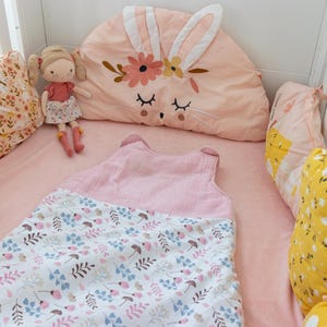 May include: A pink and white baby sleeping bag with a floral pattern. The sleeping bag is in a crib with a pink bunny-shaped pillow and yellow floral patterned pillows.
