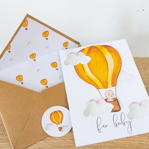 May include: A white card with a yellow and orange hot air balloon with a baby inside. The card says "for baby" and has white clouds around the balloon. The card is on a wooden surface with a brown envelope and a round sticker with a yellow and orange hot air balloon.