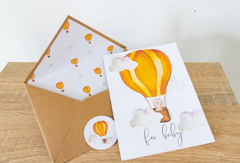 May include: A white card with a yellow and orange hot air balloon with a baby inside. The card says "for baby" and has white clouds around the balloon. The card is on a wooden surface with a brown envelope and a round sticker with a yellow and orange hot air balloon.
