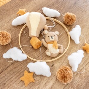 May include: A wooden hoop mobile with a brown teddy bear, a hot air balloon, clouds, and stars. The mobile is made of felt and is a neutral color palette of brown, white, and yellow.