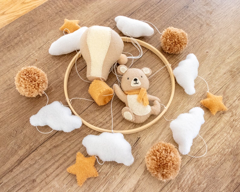 May include: A wooden hoop mobile with a brown teddy bear, a hot air balloon, clouds, and stars. The mobile is made of felt and is a neutral color palette of brown, white, and yellow.
