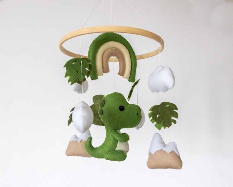 May include: A green felt dinosaur mobile with a rainbow, clouds, and palm leaves hanging from a wooden ring.