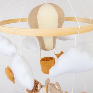 May include: A wooden baby mobile with a beige and white hot air balloon, white felt clouds, and a brown felt animal. The mobile is hanging from a wooden ring.