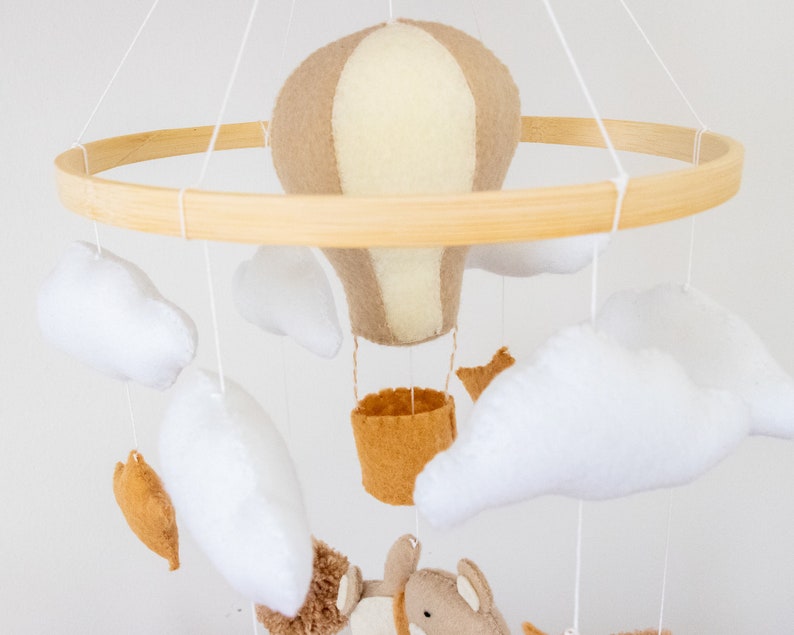 May include: A wooden baby mobile with a beige and white hot air balloon, white felt clouds, and a brown felt animal. The mobile is hanging from a wooden ring.