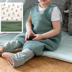 May include: A child wearing a green waffle-weave sleep sack with a white shirt underneath. The child is sitting on a light blue and white play mat with a gray elephant plush toy in the background.