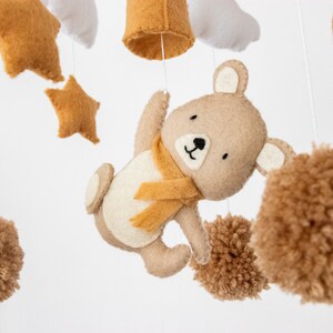 May include: A brown felt bear with a yellow scarf hangs from a mobile with brown pom-poms, stars, and clouds.