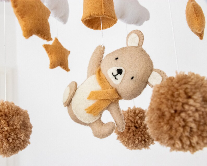 May include: A brown felt bear with a yellow scarf hangs from a mobile with brown pom-poms, stars, and clouds.
