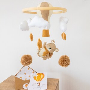 May include: A wooden baby mobile with a brown and beige hot air balloon, white clouds, brown pom poms, and a brown bear with a scarf. The mobile is hanging from a wooden ring. There is a card with a yellow hot air balloon and the text "for baby" on a wooden table.