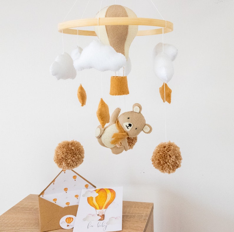 May include: A wooden baby mobile with a brown and beige hot air balloon, white clouds, brown pom poms, and a brown bear with a scarf. The mobile is hanging from a wooden ring. There is a card with a yellow hot air balloon and the text "for baby" on a wooden table.
