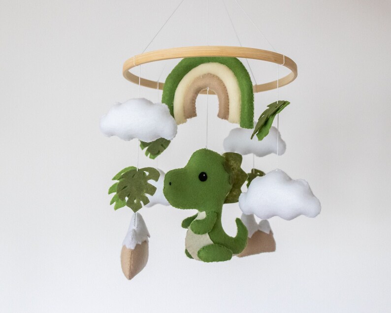 May include: A green felt dinosaur with a rainbow, clouds, and palm leaves hanging from a wooden ring. This is a baby mobile.