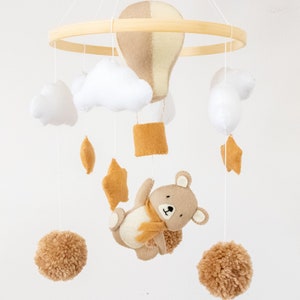 May include: A beige and yellow hot air balloon, white clouds, brown pom poms, a brown bear with a yellow scarf, and yellow stars hang from a wooden ring. This is a felt baby mobile.