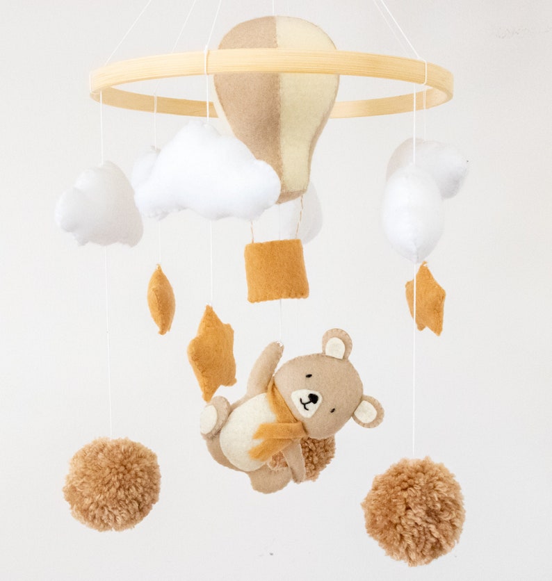 May include: A beige and yellow hot air balloon, white clouds, brown pom poms, a brown bear with a yellow scarf, and yellow stars hang from a wooden ring. This is a felt baby mobile.
