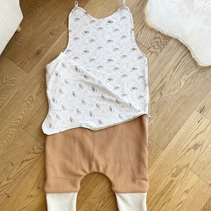 May include: A two-piece baby outfit. The top is a white bib with a pattern of brown and gray leaves and small animals. The bottom is tan waffle-knit pants with white cuffs. The outfit is laid out on a wooden floor.