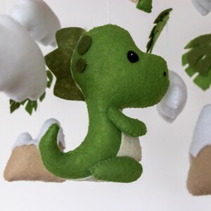 May include: A green felt dinosaur mobile hanging with white felt clouds, brown felt mountains, and green felt leaves.