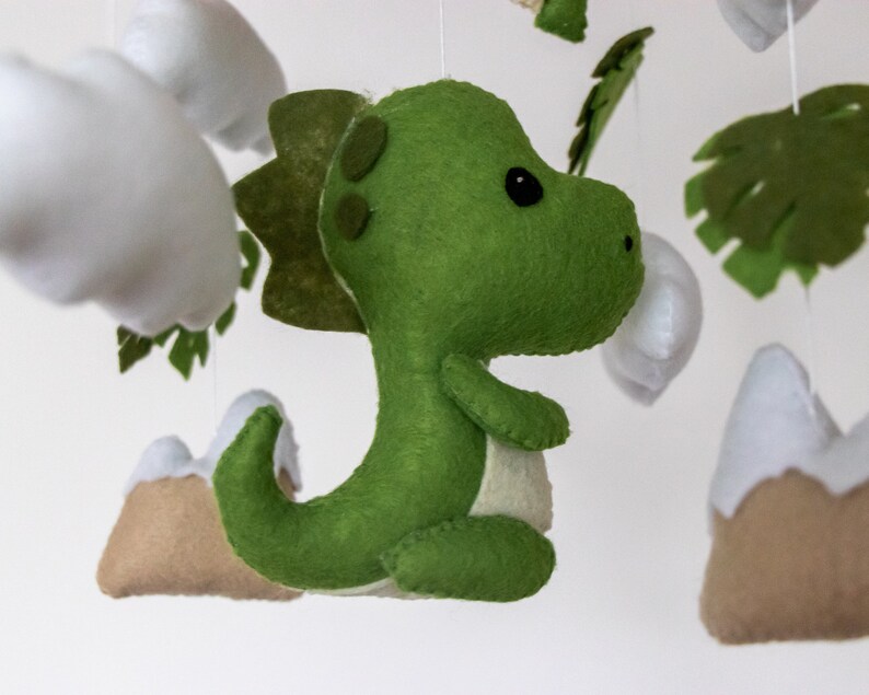May include: A green felt dinosaur mobile hanging with white felt clouds, brown felt mountains, and green felt leaves.