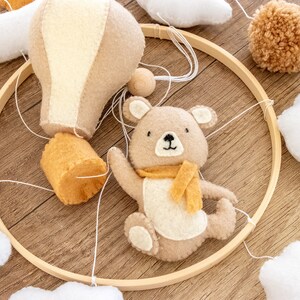 May include: A wooden mobile with a brown bear, a hot air balloon, and clouds. The mobile is made of felt and is hung from a wooden ring.
