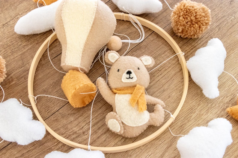 May include: A wooden mobile with a brown bear, a hot air balloon, and clouds. The mobile is made of felt and is hung from a wooden ring.