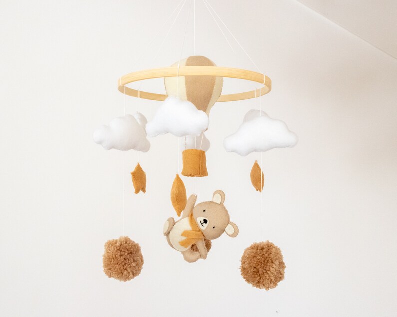 May include: A wooden baby mobile with a hot air balloon, clouds, stars, and a teddy bear. The mobile is made of felt and has a neutral color palette of white, brown, and yellow.
