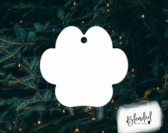 May include: White paw print shaped ornament with a hole at the top for hanging. The ornament is against a background of a green Christmas tree with lights.
