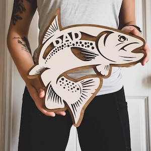 May include: A white wooden fish cutout with the word "DAD" carved into it. The fish has brown spots and a brown outline.