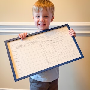 May include: A young boy holds up a white board with a blue frame. The board has a chart for tracking daily activities, a section for learning shapes, and a section for learning numbers and the alphabet. The board says "You did it! My name is LANDON."