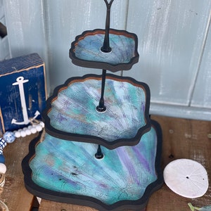 Könnte beinhalten: Ein dreistöckiger Servierständer in Form von Muscheln. Der Ständer ist in Blautönen, Grün- und Violetttönen mit einem schwarzen Rand bemalt. Der Ständer hat einen schwarzen Metallgriff.