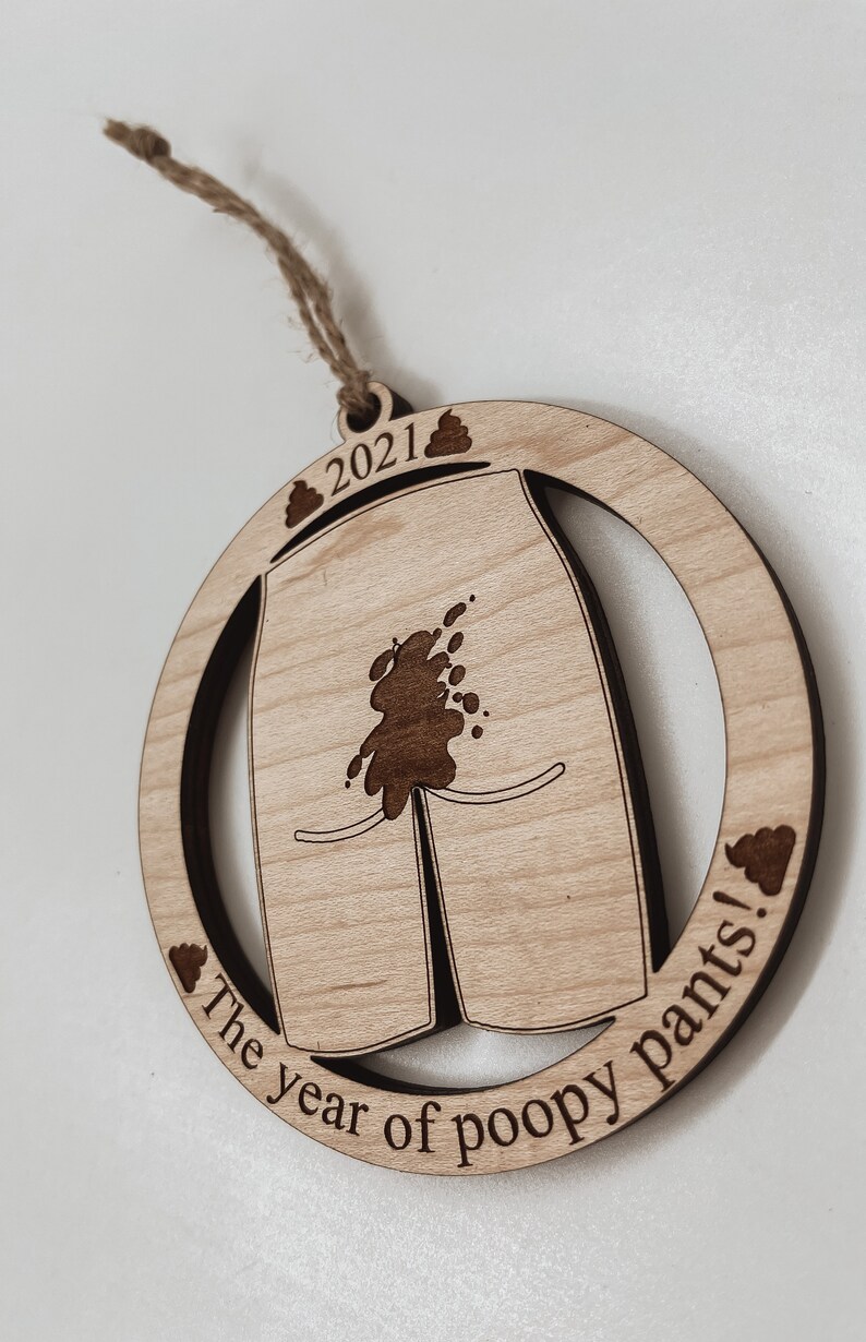 May include: A round wooden ornament with a brown stain. It features a pair of pants with a brown stain on the front and the text "The year of poopy pants!" and "2021" with a poop emoji on each side.