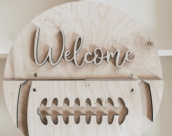 May include: A wooden round sign with the word "Welcome" cut out in script font. The sign is shaped like a football with the laces cut out.