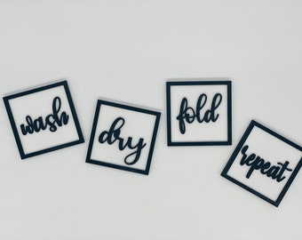 May include: Four square wooden signs with black frames and white backgrounds. Each sign has a black cursive word: "wash", "dry", "fold", and "repeat". The signs are arranged diagonally on a white surface.
