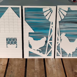 Może przedstawiać: Trzy białe drewniane dekoracje ścienne w stylu wiejskim. Pierwsza przedstawia dom z dachem i oknem z kratą imitującą drut do kur. Druga przedstawia wiatrak i kurę. Trzecia przedstawia koguta. Tło każdego obrazu to akwarela w odcieniach niebieskiego i szarego.