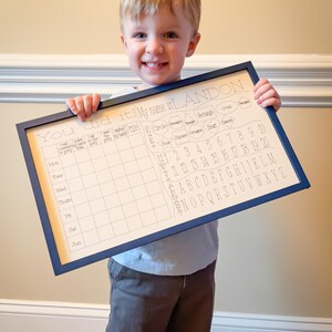 May include: A white board with a blue frame, featuring a chart for tracking daily activities and a section for learning shapes, numbers, and the alphabet. The board says "You did it! My name is LANDON". A young boy is holding the board in front of him.