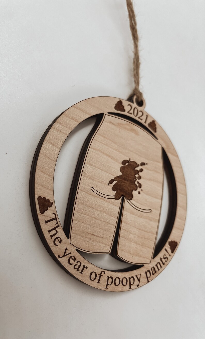 May include: A round wooden ornament with a pair of pants with a brown stain on them. The ornament is inscribed with the text "The year of poopy pants!" and "2021".