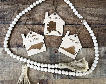Pode incluir: Três ornamentos de madeira em forma de casas com os estados do Alasca, Carolina do Norte e Virgínia gravados neles. Os ornamentos estão pendurados em uma guirlanda de contas brancas com uma borla marrom.