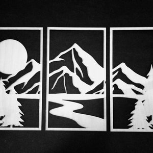 Può includere: Un'opera d'arte murale in tre pannelli con sfondo nero e design ritagliato in bianco. Il design presenta una catena montuosa, un fiume, una luna piena e alberi.
