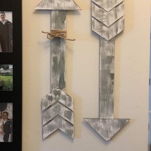 May include: Two wooden arrows with a distressed white and gray paint finish. The arrows are pointing in opposite directions. The arrow pointing up has a brown twine bow tied around the shaft.