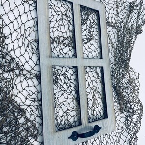 以下が含まれることがあります： 4 つのガラス窓が付いた、風化した灰色の木製の窓枠。枠には黒い金属製の取っ手が付いており、茶色の漁網を背景に置かれています。