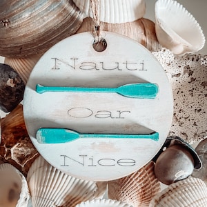 May include: A white wooden round ornament with the text "Nauti Oar Nice" and two teal blue painted oars. The ornament is surrounded by seashells.