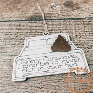 May include: A wooden Christmas ornament shaped like a bed frame, with a brown poop emoji attached. The ornament has the text "Merry Christmas! I promise to never shit in your bed."