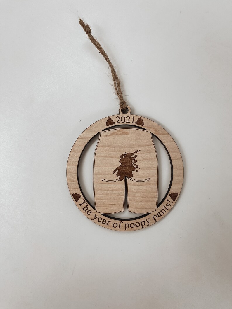 May include: A wooden Christmas ornament with a brown poop stain on a pair of white pants. The ornament is round and has the text "2021 The year of poopy pants!" on it.