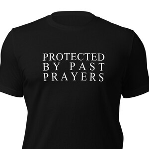 Op de afbeelding: Zwart T-shirt met witte tekst die "PROTECTED BY PAST PRAYERS" luidt.