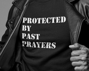 Puede incluir: Una foto en blanco y negro de una persona que lleva una chaqueta de cuero negra sobre una camiseta negra con el texto "PROTECTED BY PAST PRAYERS" impreso en blanco. La persona lleva un sombrero negro y tiene barba.
