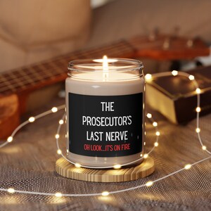 May include: A brown candle with a black label that reads "The Prosecutor's Last Nerve - Oh Look, It's On Fire". The candle is lit and sitting on a wooden coaster.