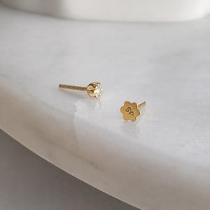 May include: Two gold stud earrings. One earring features a small, clear gemstone. The other earring is a flower shape with the letters "SP" engraved on it. The earrings are on a white marble surface.