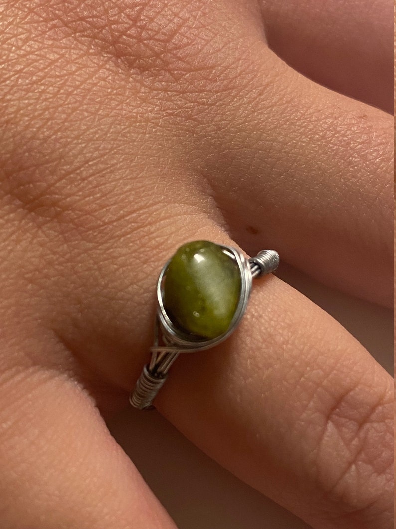 May include: A silver wire wrapped ring with a green gemstone. The ring is on a finger.