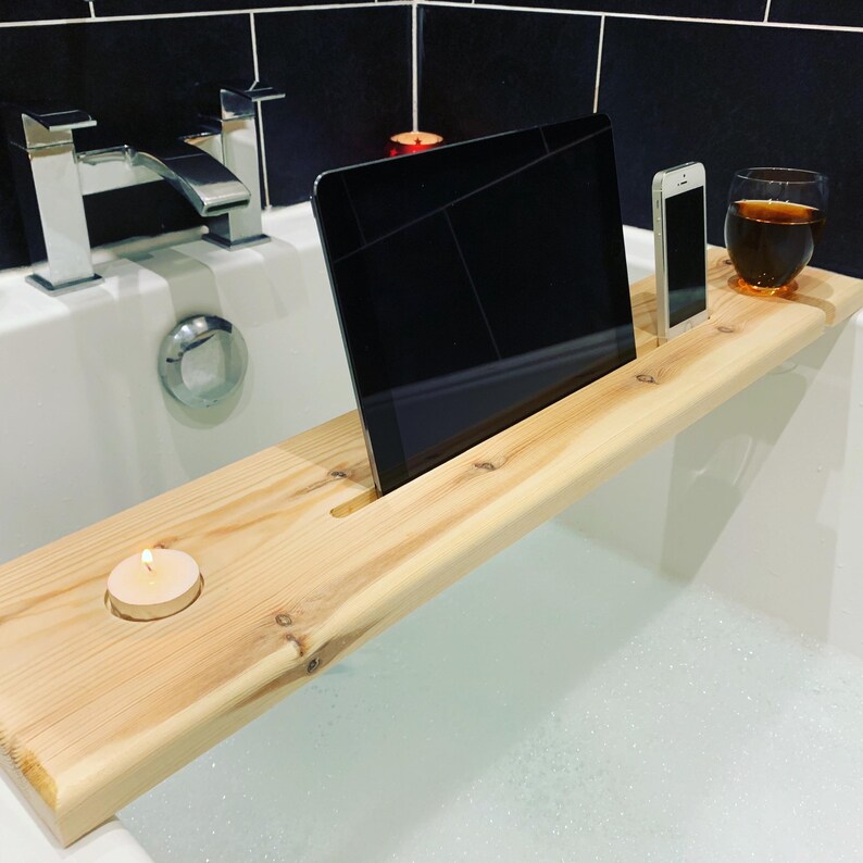 May include: A wooden bath tray with a slot for a tablet, a phone holder, and a small candle holder. The tray is made of natural wood and has a light brown colour.