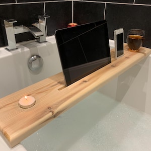 May include: A wooden bath tray with a slot for a tablet, a phone holder, and a space for a candle. The tray is in a bathtub filled with bubbles.